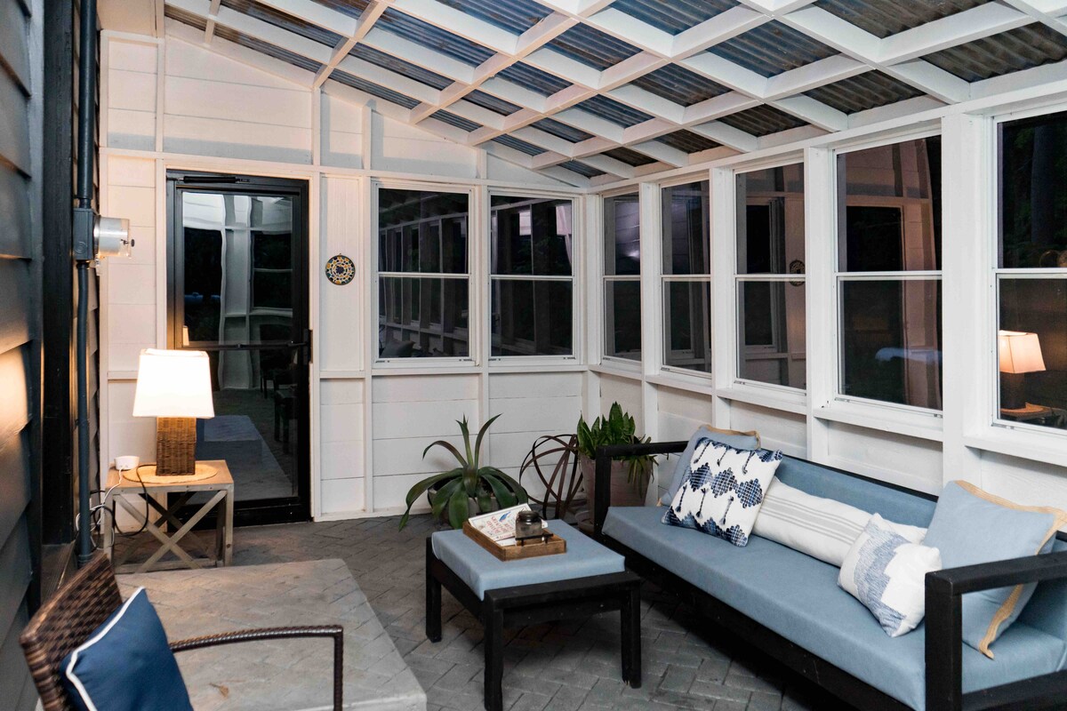 A newly renovated sunroom features large windows allowing abundant natural light. Comfortable seating is arranged around a wooden coffee table. A lamp provides soft lighting, and potted plants add a touch of greenery, enhancing the welcoming ambiance.