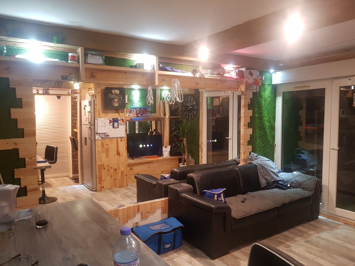 A spacious living area showcases a large black sofa accompanied by throw blankets. Walls feature wooden accents and greenery. A flat-screen television is visible, surrounded by decorative elements. Natural light streams through large glass doors, enhancing the open and inviting feel of the space.