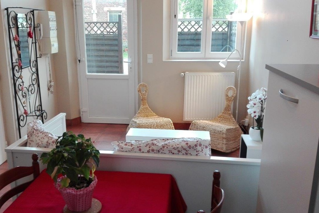 A cozy living area is seen, featuring two rattan chairs and a small table. Natural light enters through the windows, highlighting the warm tones of the decor. A plant sits on the table, adding a touch of greenery to the space.