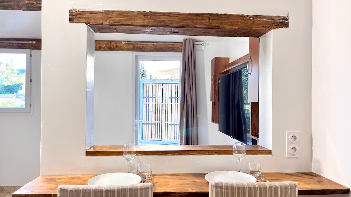 A minimalist kitchenette is showcased with a wooden countertop, featuring two place settings and glasses. A window with light curtains allows natural light to enter, creating a bright atmosphere. A wall-mounted television is visible in the background, adding functionality to the space.