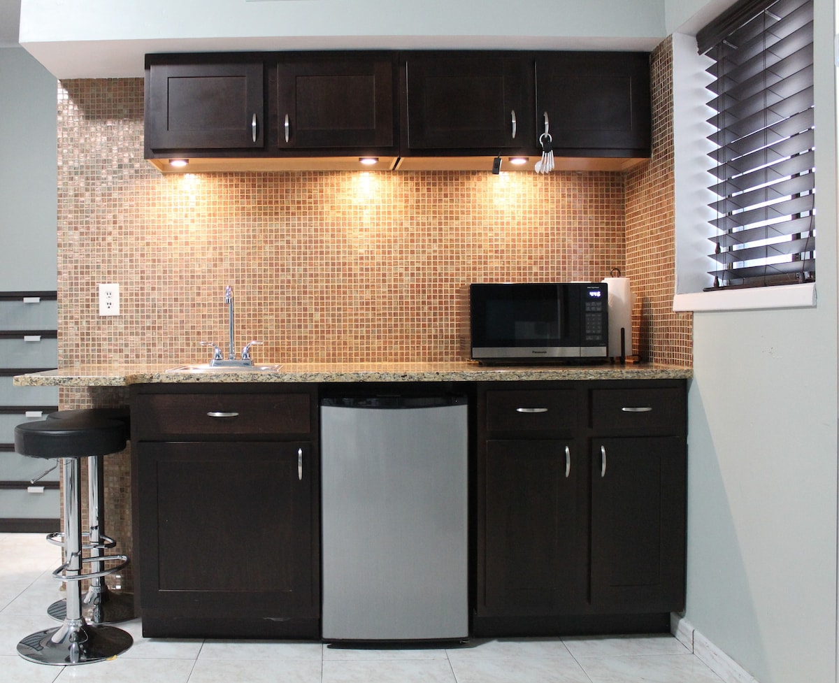 A modern kitchenette includes dark cabinetry and a beige mosaic tile backsplash. Appliances feature a stainless steel refrigerator, microwave, and a sleek sink. Under-cabinet lighting allows for a bright workspace, complemented by a small bar area with a black stool.