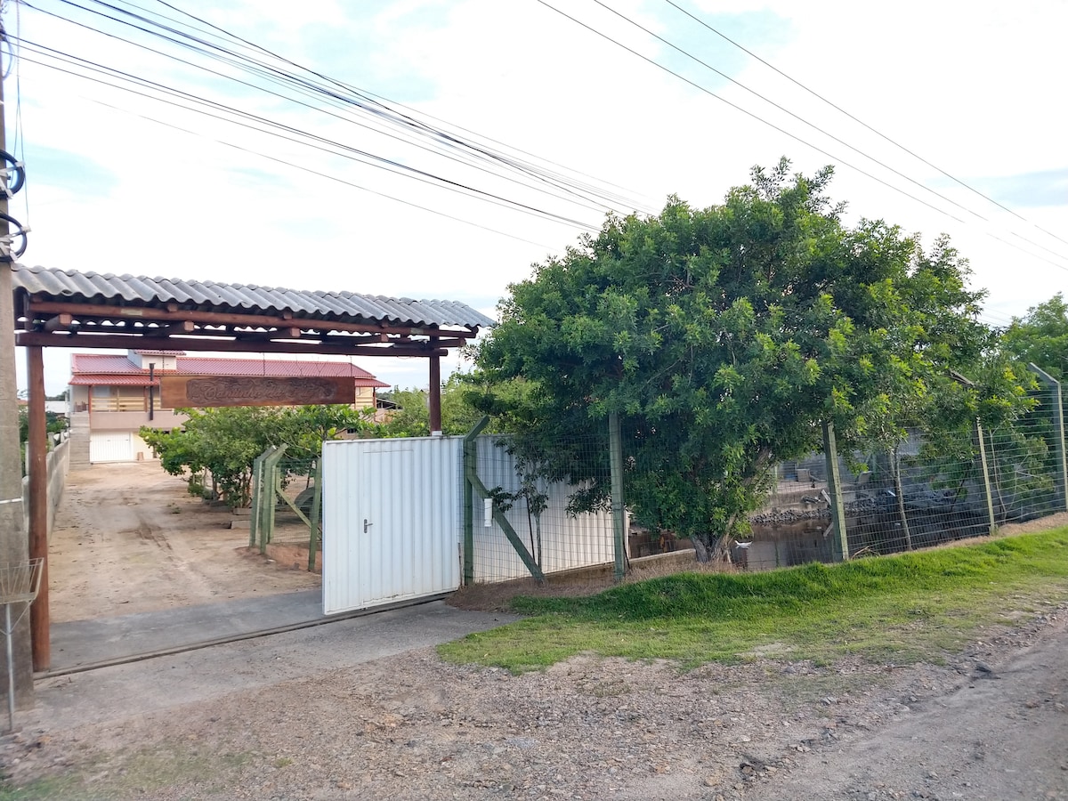 An entrance gate leads into a spacious outdoor area, bordered by a fence and greenery. A gravel pathway is seen, along with a large tree providing shade. The scene is complemented by a clear sky and power lines in the background.