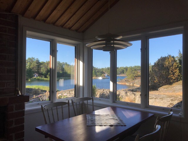 Large windows frame a serene view of the water and surrounding nature, allowing ample natural light to fill the dining area. A wooden table sits at the center, complemented by several chairs. The calming landscape of trees and rocky shores can be seen in the distance.