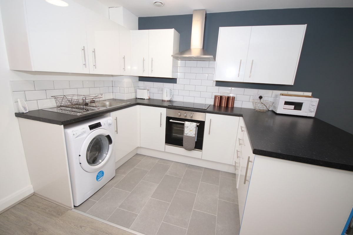 A modern kitchen is equipped with white cabinetry and gray tiled flooring. An electric stove and microwave are visible, alongside a washing machine. The kitchen features a sleek countertop and a modest amount of dishware, emphasizing a functional and tidy cooking environment.