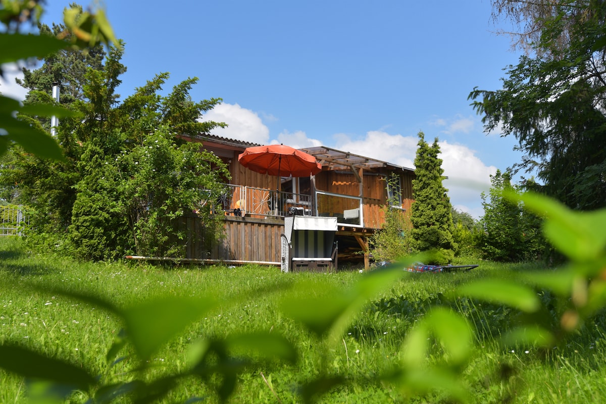 A charming wooden holiday house is set amidst lush greenery, with a vibrant orange umbrella providing shade on the deck. The building features a rustic exterior, and the surrounding grass is dotted with wildflowers, enhancing the serene outdoor environment.
