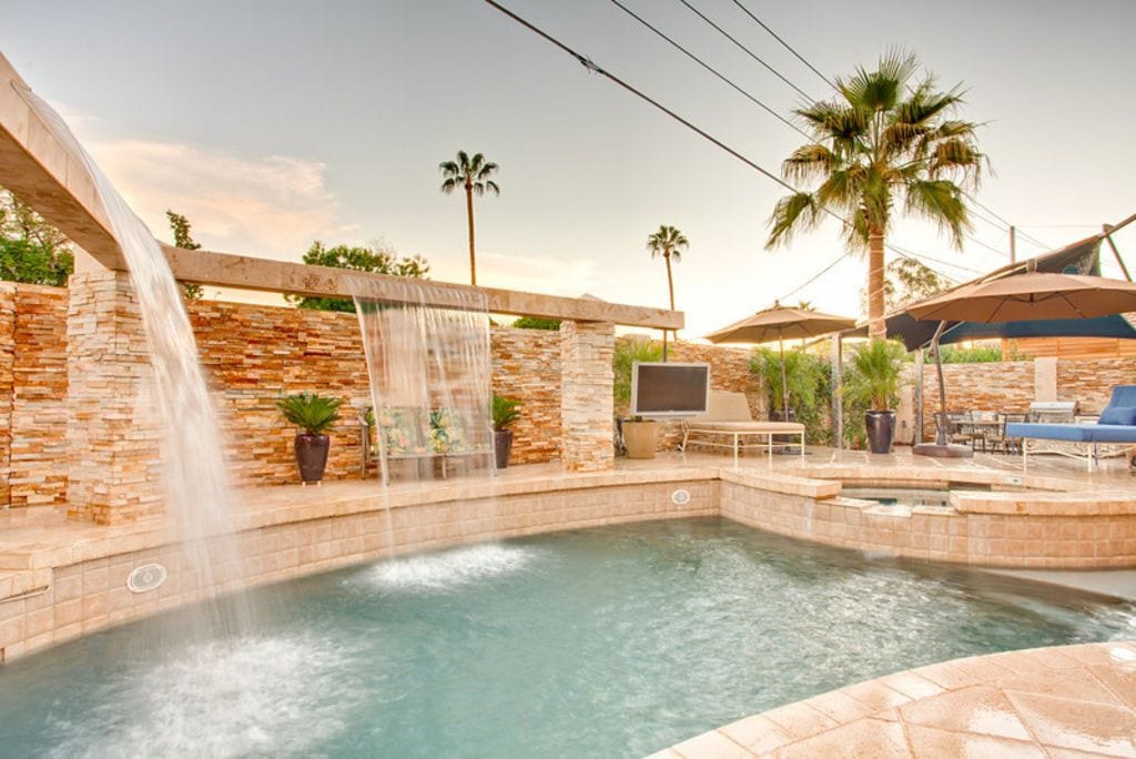 A backyard pool area is highlighted by water cascading from a stone feature, surrounded by palm trees and vibrant greenery. loungers and umbrellas offer shaded seating, while a TV is mounted on a wall for entertainment. The gentle curves of the pool invite relaxation.