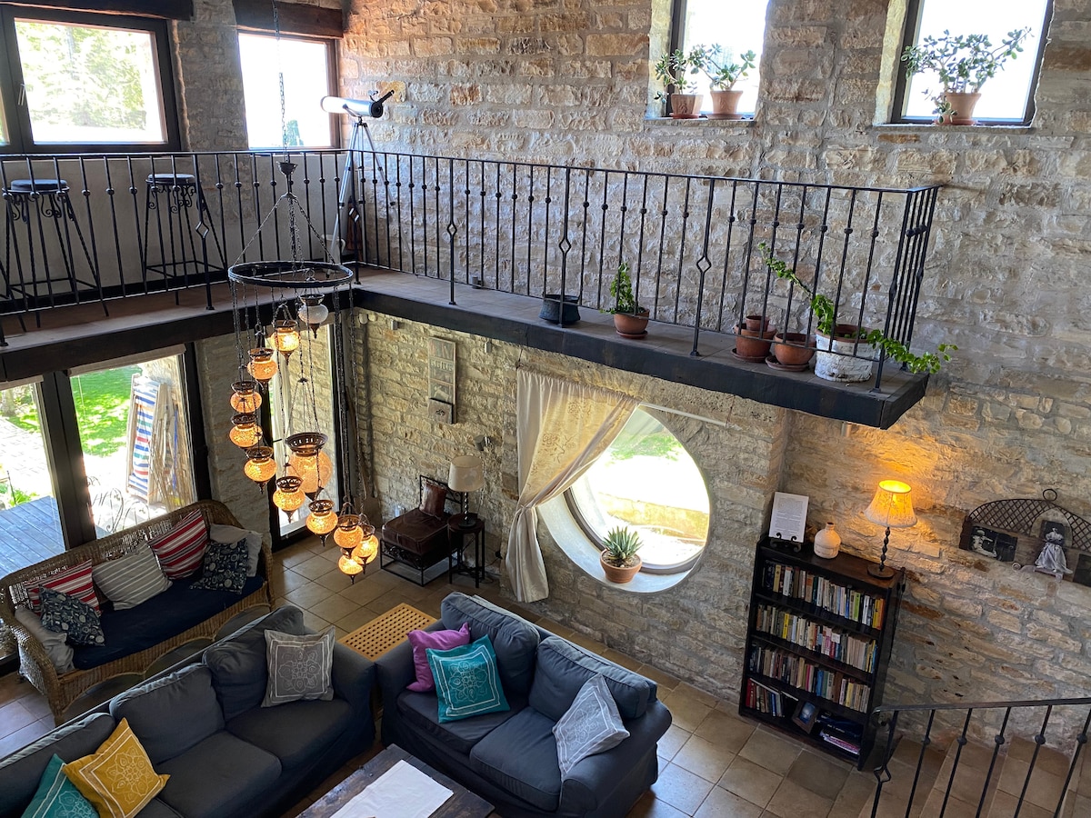 A spacious living room is highlighted by high stone walls and large round windows allowing natural light to fill the space. A balcony with potted plants overlooks the area, while a cozy seating arrangement is complemented by colorful throw pillows. A chandelier made of multiple hanging lights adds to the ambiance.