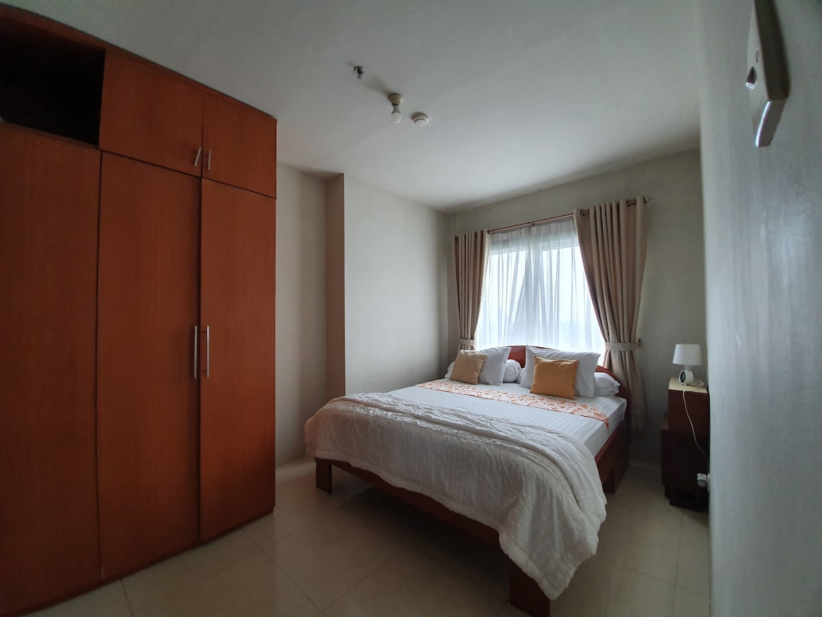 A spacious bedroom features a large bed with decorative pillows, complemented by soft linen. A wardrobe occupies one side, while a bedside table holds a lamp. Natural light enters through a large window adorned with light curtains.