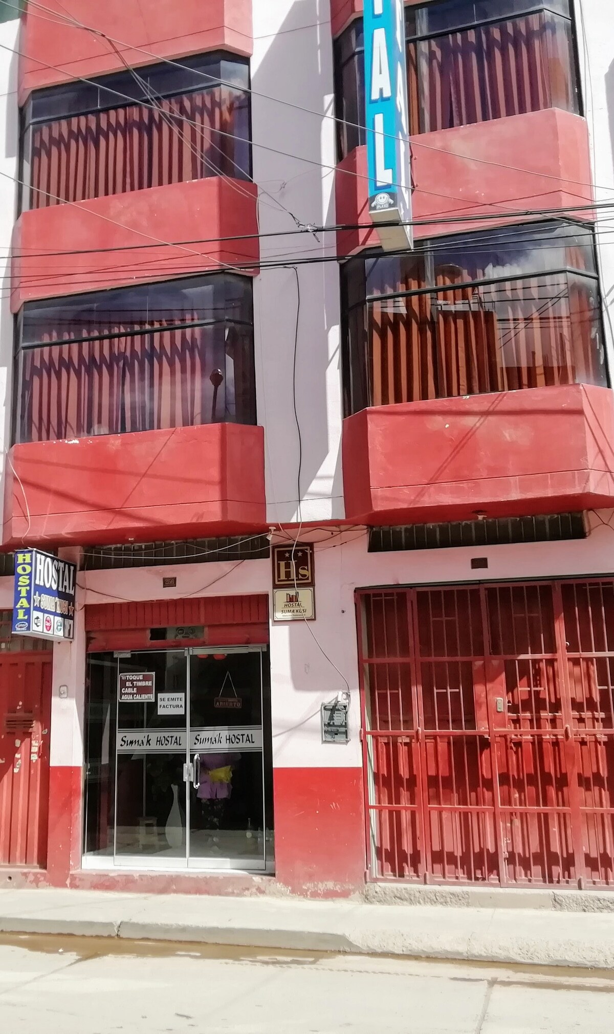The exterior of the building displays a two-story structure painted in bright red, featuring large windows with vertical blinds. A sign indicating the hostel name is prominently displayed above the entrance, which is framed by a decorative iron gate.