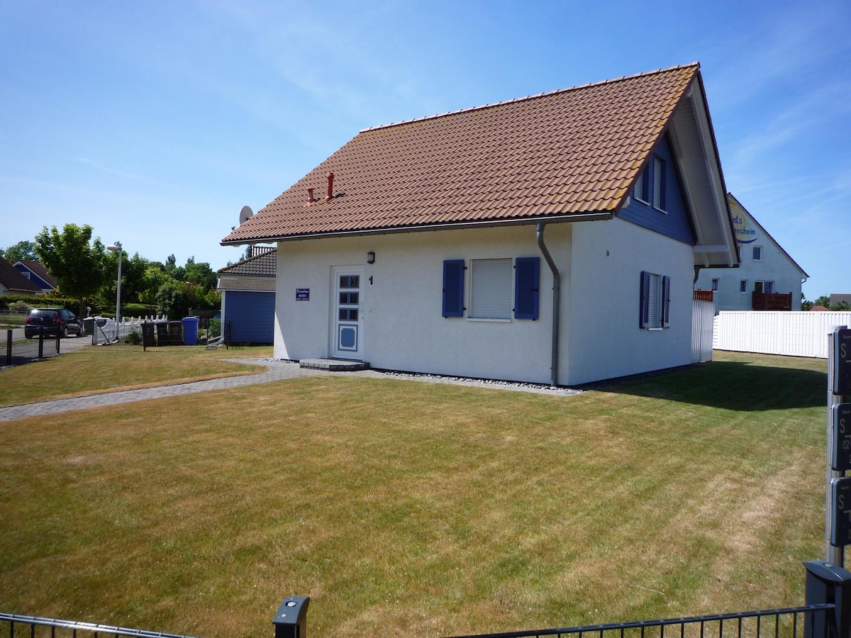 The exterior of a two-story house is depicted, featuring a sloped roof and blue shutters. A neat lawn surrounds the house, which stands alone in a spacious area. A gravel pathway leads to the front door, and nearby, a fence and trees are visible.