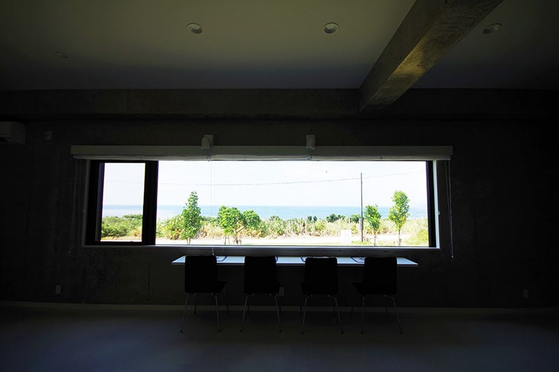 A spacious room is highlighted by large windows that provide a panoramic view of the ocean and greenery. The modern design includes a simple table with four chairs positioned beneath the windows, allowing natural light to fill the area.