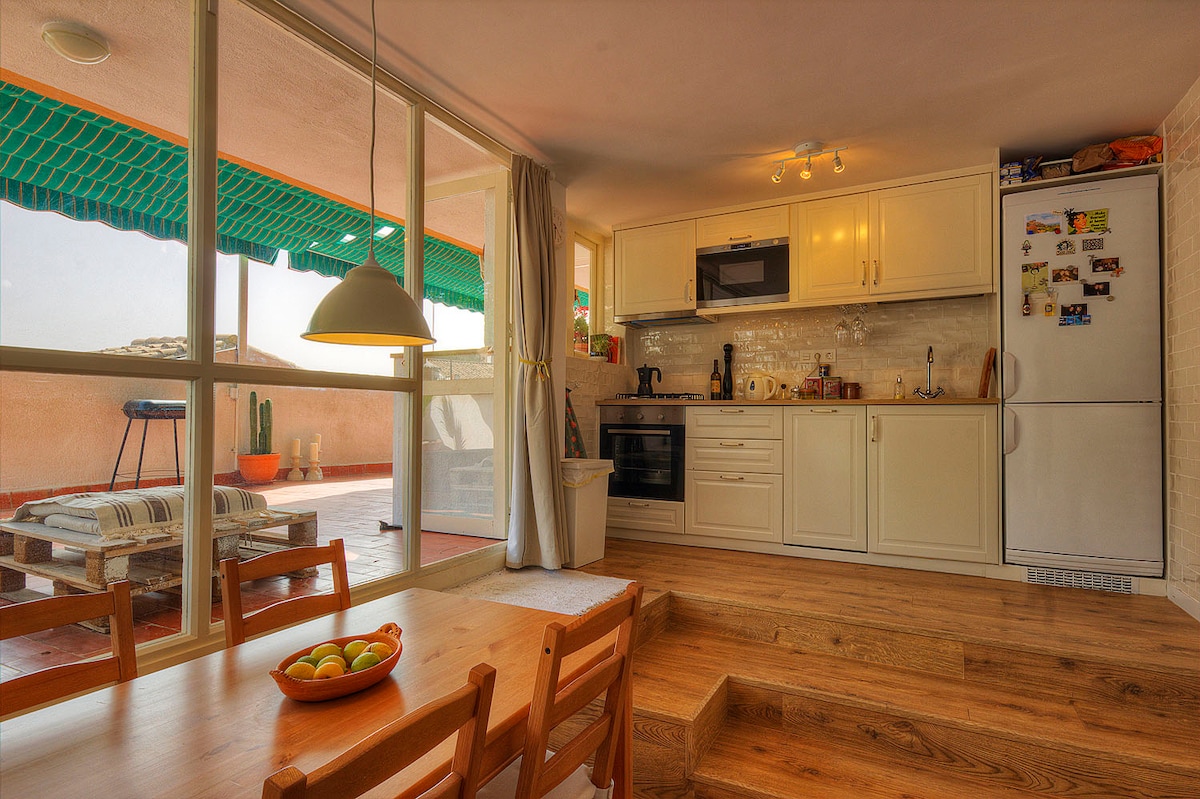A modern kitchen and dining area blend into one another, featuring white cabinetry and a wooden dining table equipped with chairs. Natural light floods the space through large windows, while a charming terrace with lounge chairs can be seen beyond the glass doors.