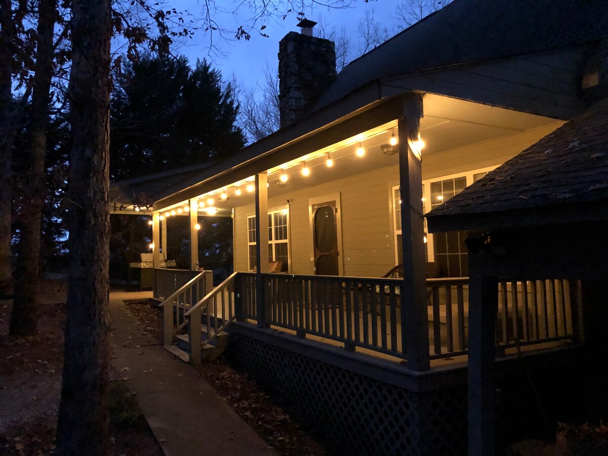A cozy porch area is illuminated by soft string lights, creating a welcoming scene during twilight. The wooden steps lead to a spacious entryway, framed by large windows that reflect the gentle evening light.