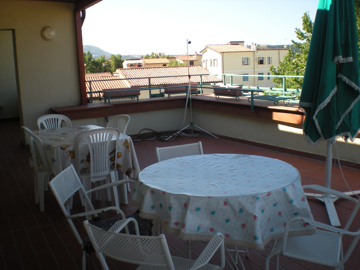 An outdoor terrace features a round table with a floral tablecloth surrounded by several white chairs. A large green umbrella stands nearby, providing shade. The area offers views of the surrounding buildings and distant hills under clear blue skies.