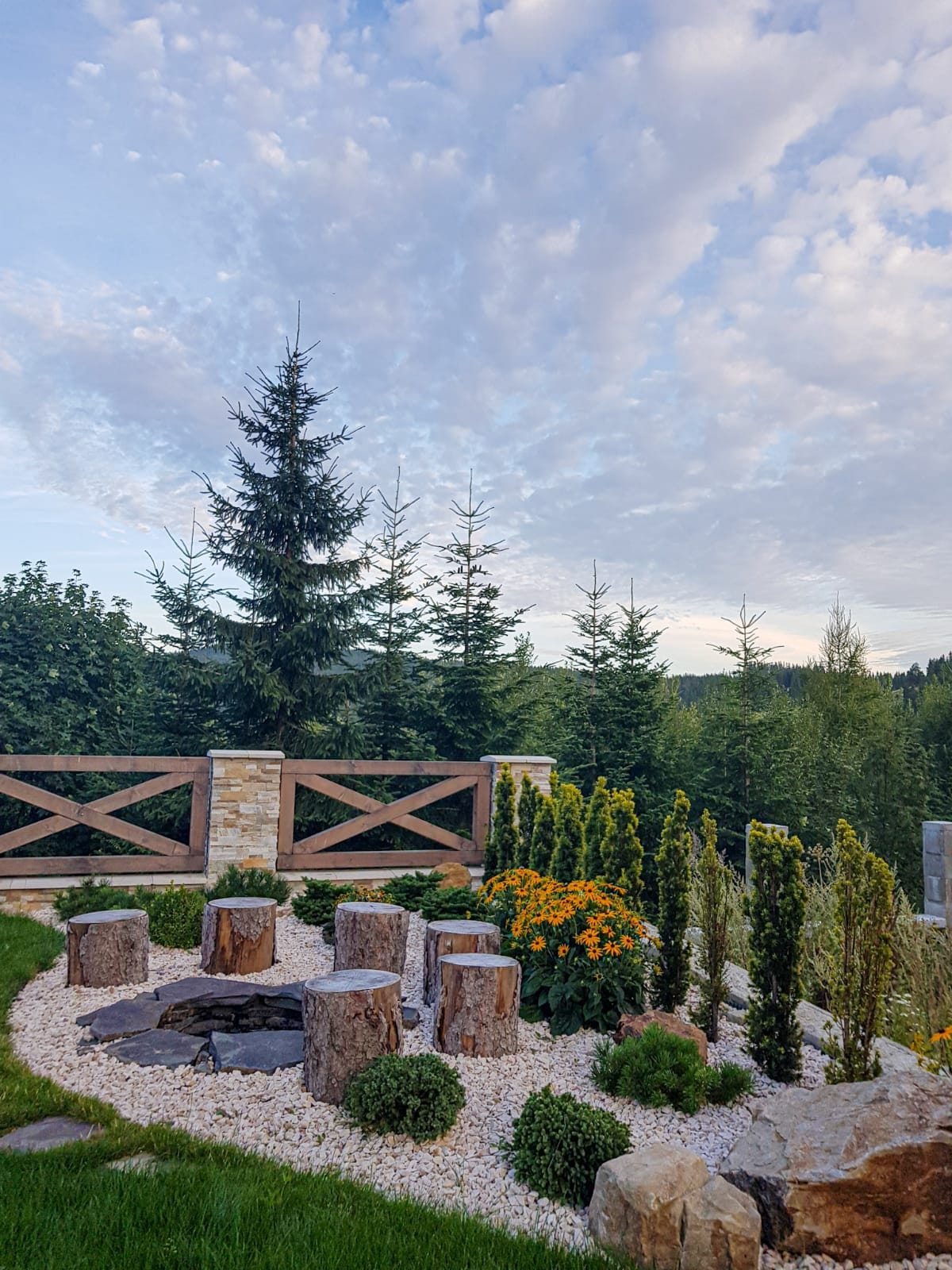 A landscaped outdoor area features natural stone seating arranged around a fire pit. Lush greenery, including tall trees and flowering plants, complements the space, while a wooden fence provides a rustic backdrop against a serene sky.
