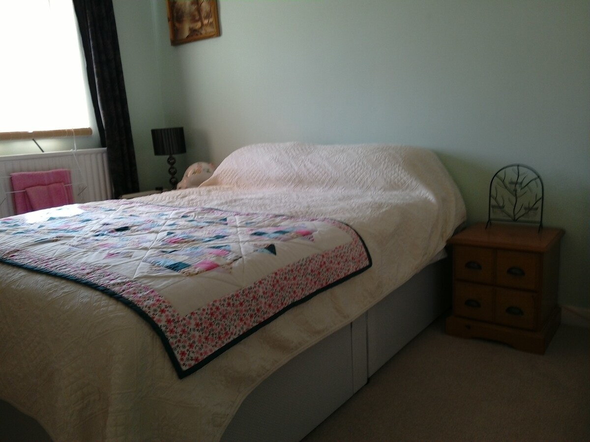 A spacious king size bed is adorned with a quilt featuring colorful patterns. A bedside table is positioned next to the bed, showcasing a decorative lamp. Natural light filters through a nearby window, partially covered by dark curtains.