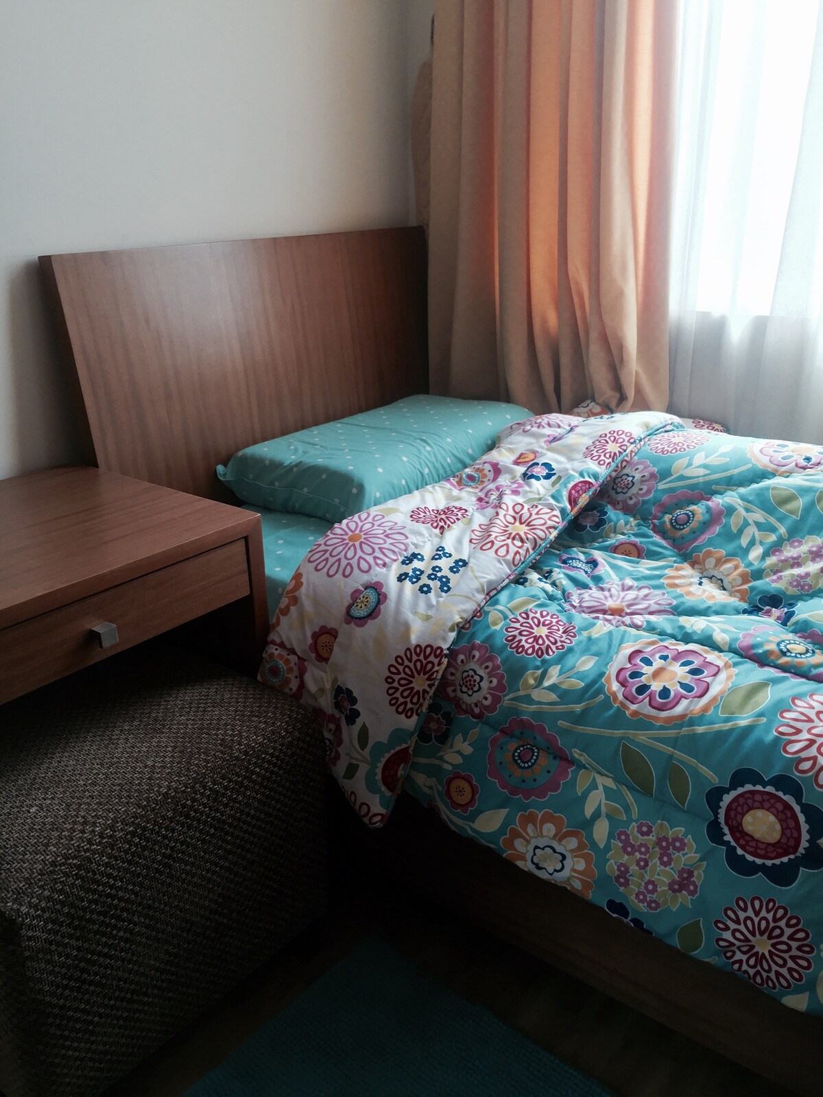 A cozy sleeping area features a wooden headboard and a spacious bed dressed in a floral comforter. A bedside table provides storage, while a soft rug lies on the floor. Natural light filters through curtains, creating a warm ambiance.
