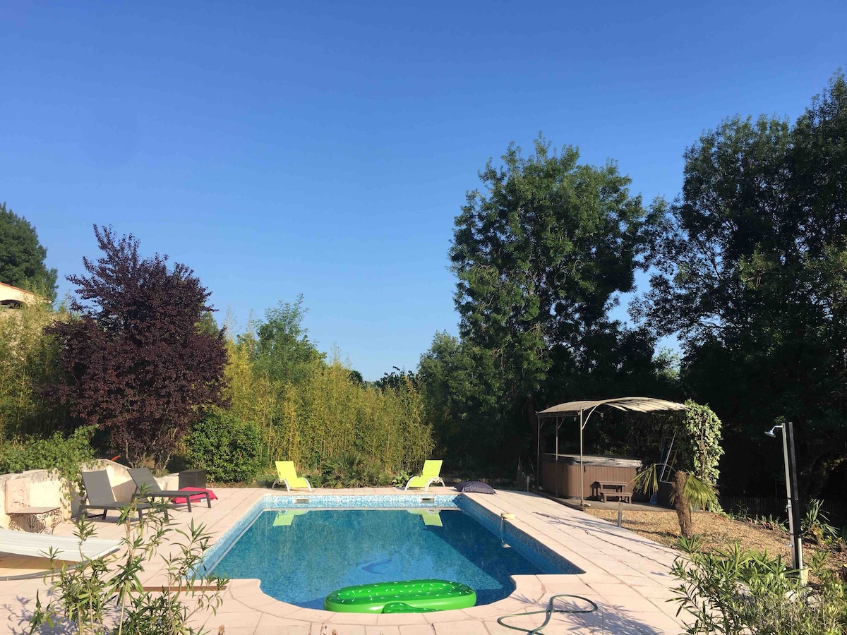 A swimming pool is surrounded by lush greenery and positioned under a clear blue sky. Two bright lounge chairs in yellow are set alongside the pool, complemented by a shaded seating area nearby. The serene landscape provides an inviting outdoor space for relaxation.