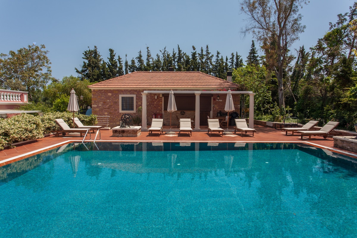 A spacious pool area is surrounded by neatly arranged lounge chairs under shaded umbrellas. The inviting water reflects the bright blue sky, while lush greenery frames the rustic stone house in the background, creating a serene outdoor setting.