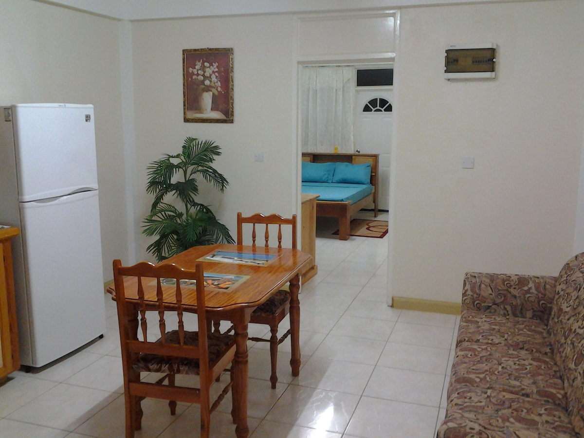 A dining area features a wooden table with four chairs, complemented by a patterned couch in a neutral tone. A refrigerator is positioned to the side, with light-colored tiles enhancing the space. A doorway leads to a brightly colored sleeping area in the background.