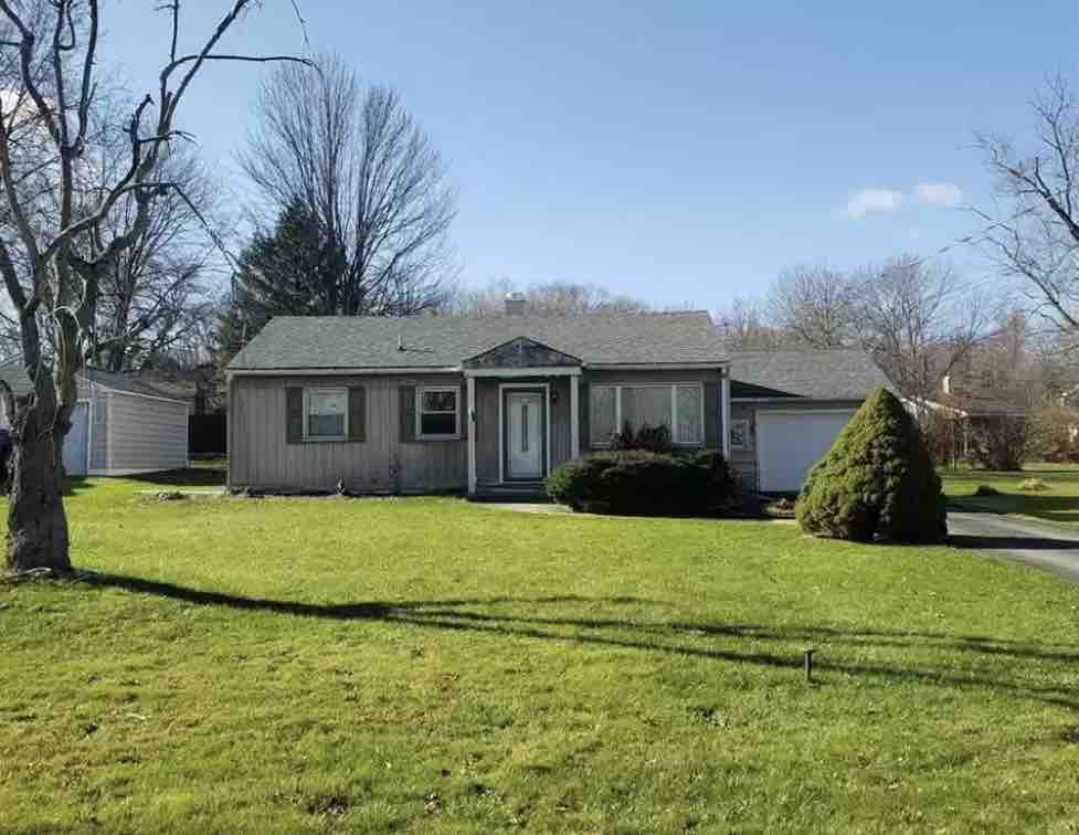 A single-story house is situated on a grassy lot, characterized by a front porch and a well-maintained exterior. Leafless trees provide a backdrop, while a manicured shrub stands near the entrance. A driveway leads to a garage on the right side of the property.