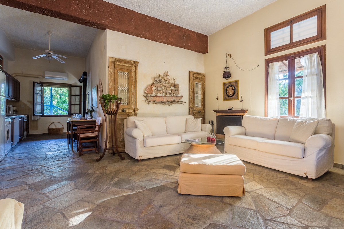 An open-plan living area features comfortable couches in neutral tones, set on a stone floor. Natural light fills the room through multiple windows, and a dining area is visible in the background, accompanied by rustic decor elements including a sailing ship artwork.