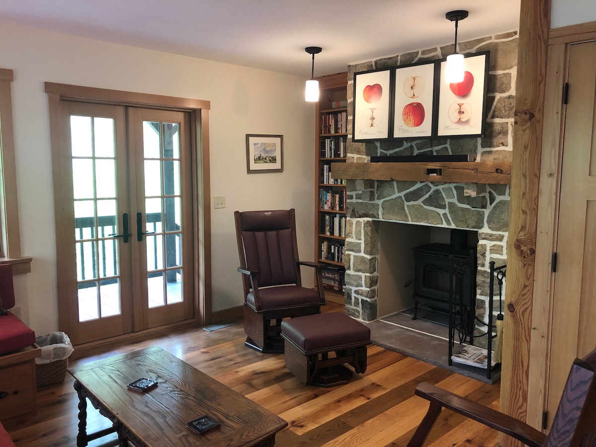 A cozy living area features a stone fireplace with a wood stove, surrounded by warm wooden accents. A comfortable rocking chair and ottoman are positioned near a wooden coffee table. The room is brightened by natural light through French doors leading to an outdoor space.