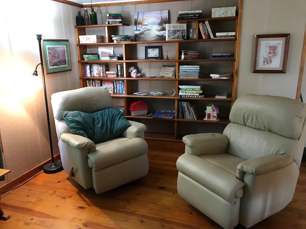 Two comfortable recliners are positioned in a cozy area, accompanied by a floor lamp. A bookshelf filled with a variety of books and decor items occupies the wall behind, adding a homely touch. Natural light brightens the warm wooden flooring.
