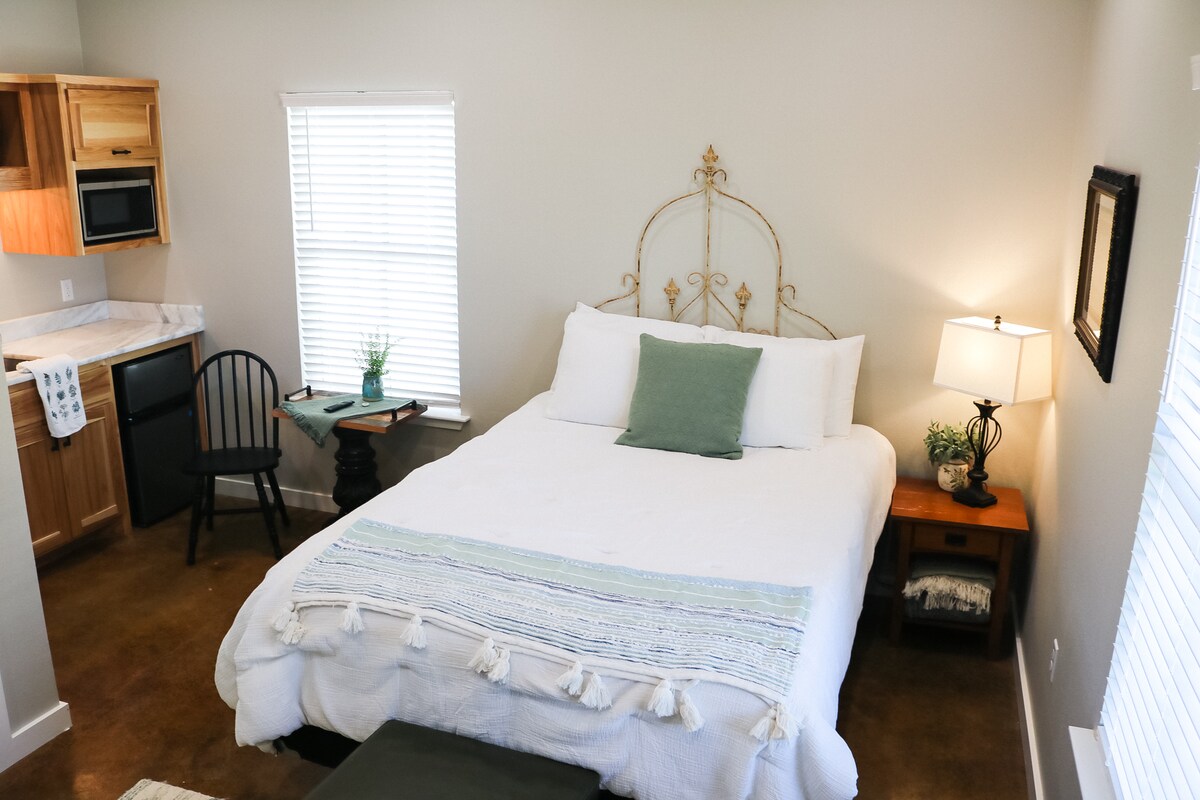 A well-lit studio features a spacious bed with a decorative headboard and a light-colored blanket. A kitchenette area is visible with wooden cabinets and a small table set for two. Natural light streams in through the window, complemented by a bedside lamp.