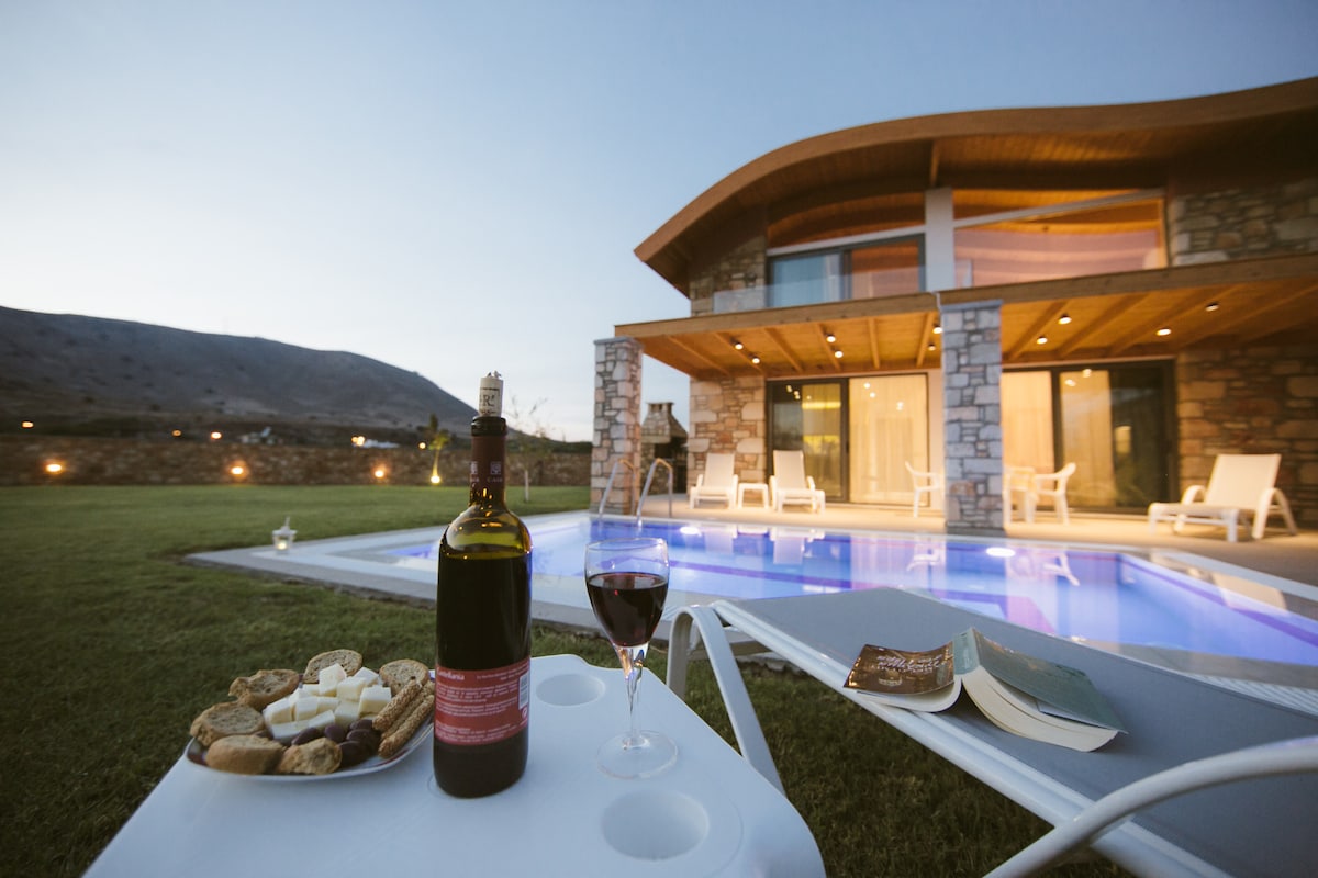 A private terrace features a swimming pool alongside modern lounge chairs. A table is set with a bottle of red wine and glasses, accompanied by an assortment of snacks. The villa's exterior showcases stone elements and expansive glass doors, framed by a mountain backdrop.