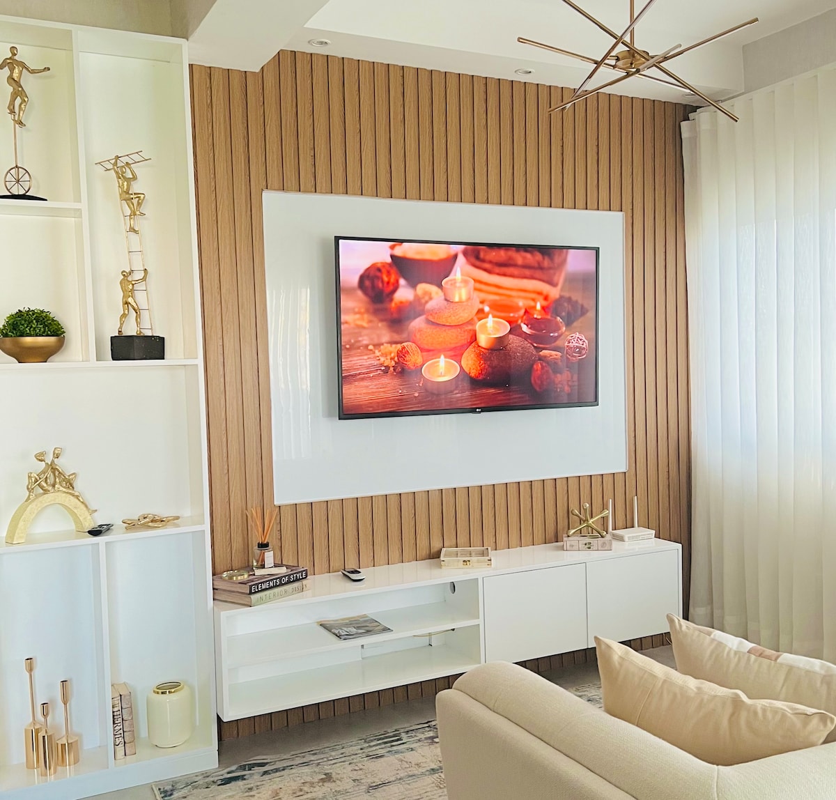 A modern living area is showcased, featuring a wall-mounted television displaying a serene image. Light-colored cabinetry with decorative accents is set against a textured wood wall, while natural light filters through sheer curtains. A neutral-toned sofa with decorative cushions complements the space.