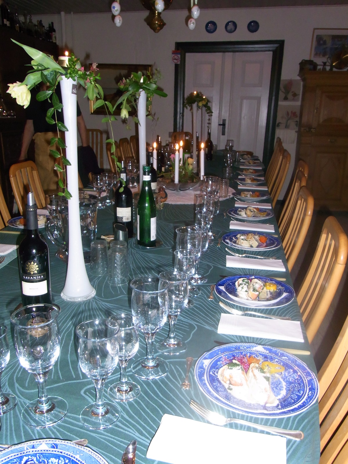 A long dining table is set for a gathering, featuring an array of glasses and elegant plates. Tall white candle holders with flowers accentuate the center. Various bottles of wine are placed along the table, creating a welcoming atmosphere for mealtime.