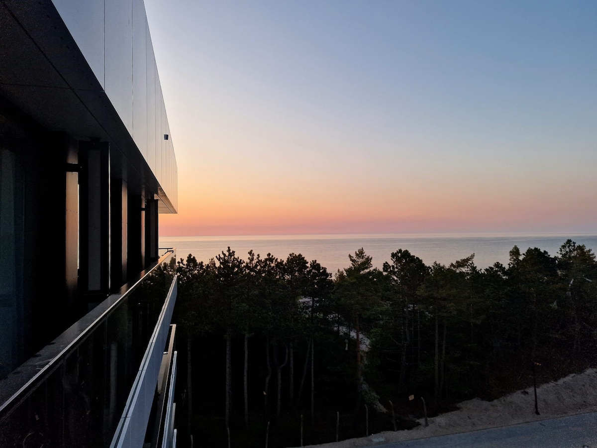 A serene view from the balcony captures a soft gradient of colors in the sky during sunset. The calm sea extends towards the horizon, surrounded by silhouettes of trees that frame the scene, offering a tranquil atmosphere.