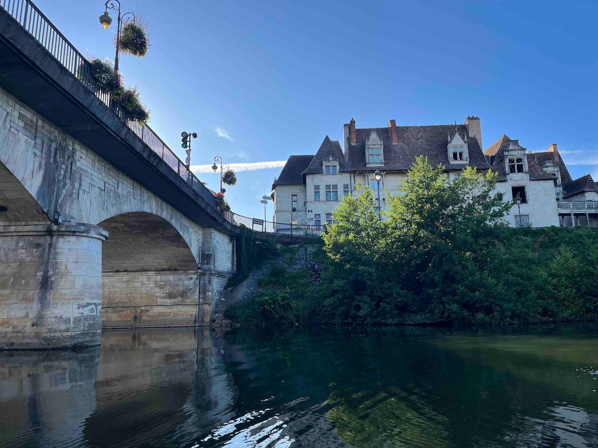 A serene riverside view is captured, showcasing a historic building alongside a stone bridge. Lush greenery and hanging flower baskets contribute to the tranquil setting, while reflections on the water create a sense of calm. The blue sky enhances the peaceful atmosphere.