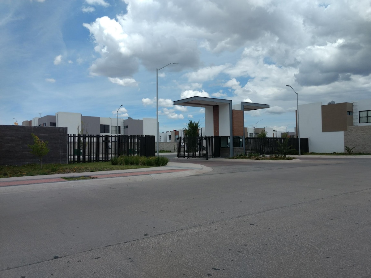A gated entrance is shown with a modern design, featuring a tall, open structure and a decorative fence. Surrounding greenery and neatly arranged pathways create a welcoming approach. The sky is partly cloudy, enhancing the clean lines of the contemporary architecture in the background.