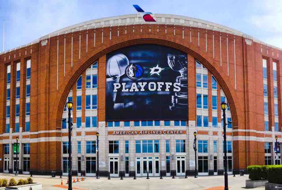 The exterior of the American Airlines Center is captured, showcasing its distinctive brick architecture. A large digital sign displays the word 'PLAYOFFS,' signifying an event, while the venue's entrance features multiple glass doors and a prominent logo banner above.