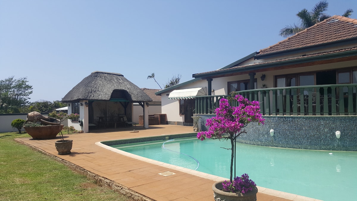 A spacious outdoor area features a swimming pool surrounded by a green lawn, with a thatched gazebo in the background. Potted plants with vibrant flowers add color to the scene, while a patio can be seen attached to the building, offering outdoor seating options.
