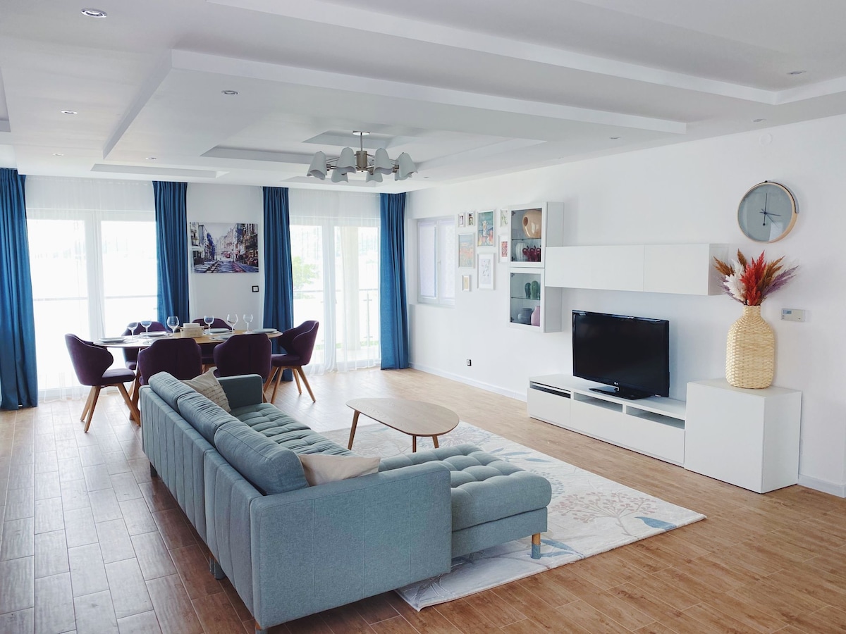 A modern living area features a light blue sectional sofa, a wooden coffee table, and a flat-screen TV on a white entertainment unit. Large windows dressed in blue curtains allow natural light to illuminate the space. A dining table is set nearby with upholstered chairs.