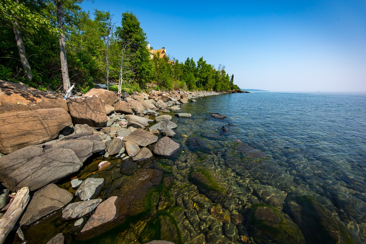 The rocky shoreline of Lake Superior features clear waters revealing colorful pebbles beneath the surface. Lush greenery lines the bank, and the horizon is visible in the distance under a bright blue sky. The natural landscape offers a serene and inviting atmosphere.