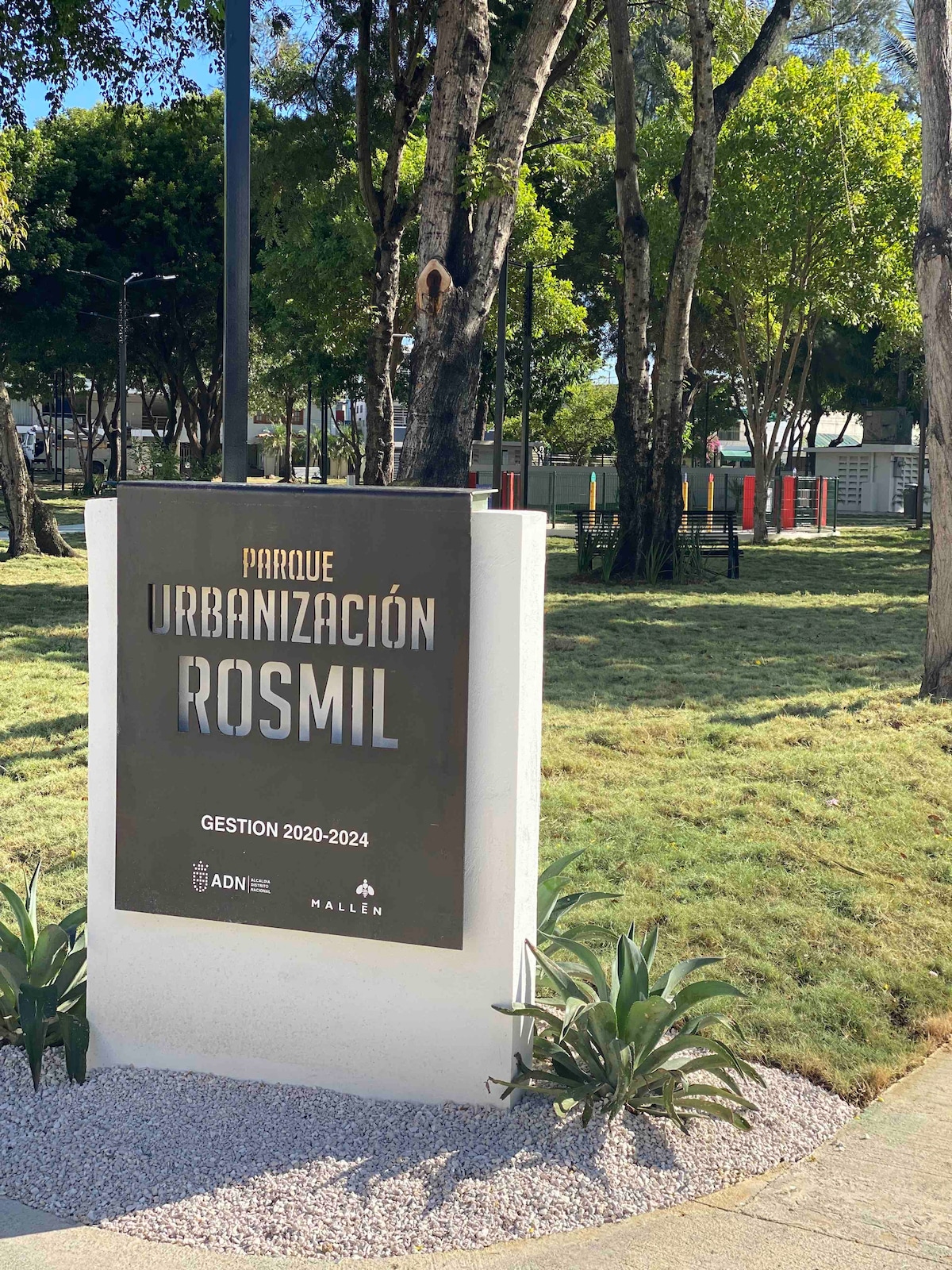 A prominent sign is displayed at the entrance of Rosmil Park, indicating the park's name and management information. Surrounding greenery includes well-maintained grass and landscaping elements, contributing to a peaceful outdoor environment. Tall trees provide shade in the background.