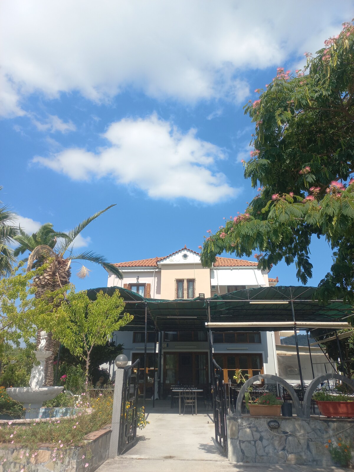 A charming house is set against a bright blue sky, framed by lush green trees. A spacious patio with tables is visible in the foreground, complemented by tropical plants and colorful floral arrangements. The building features a light facade with balconies and a classic tiled roof.