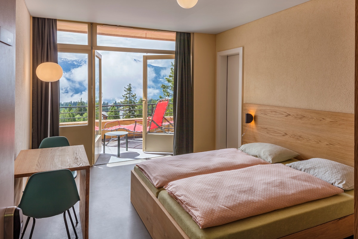 A bright room features a spacious bed with pale bedding and a wooden frame. A table and chairs are positioned near the tall windows, which open to a balcony offering scenic views. Soft grey walls complement the natural light flooding the space.