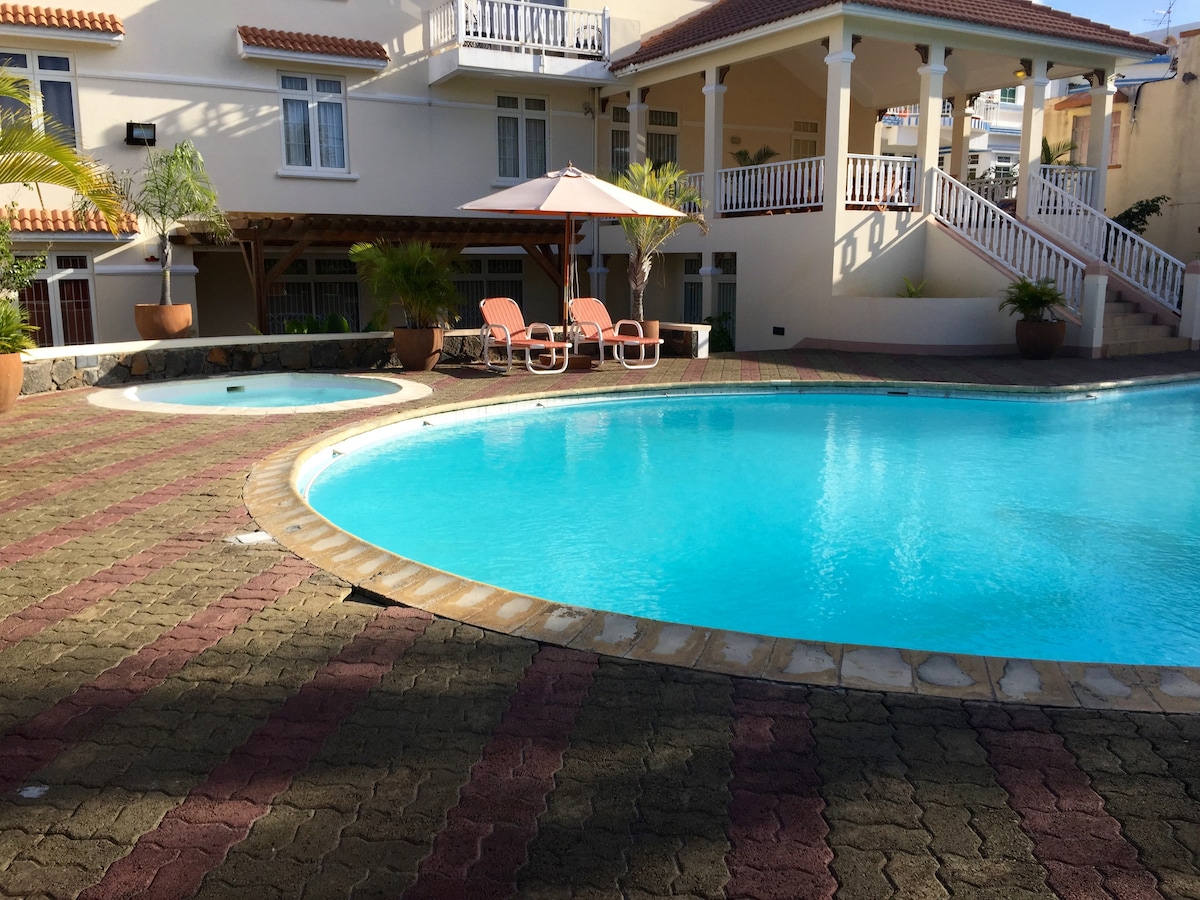 A large swimming pool area is visible with two spacious pools. Sun loungers with an umbrella are positioned beside the pools, surrounded by landscaped greenery. The well-maintained courtyard features patterned stone pathways, contributing to a relaxing outdoor environment.