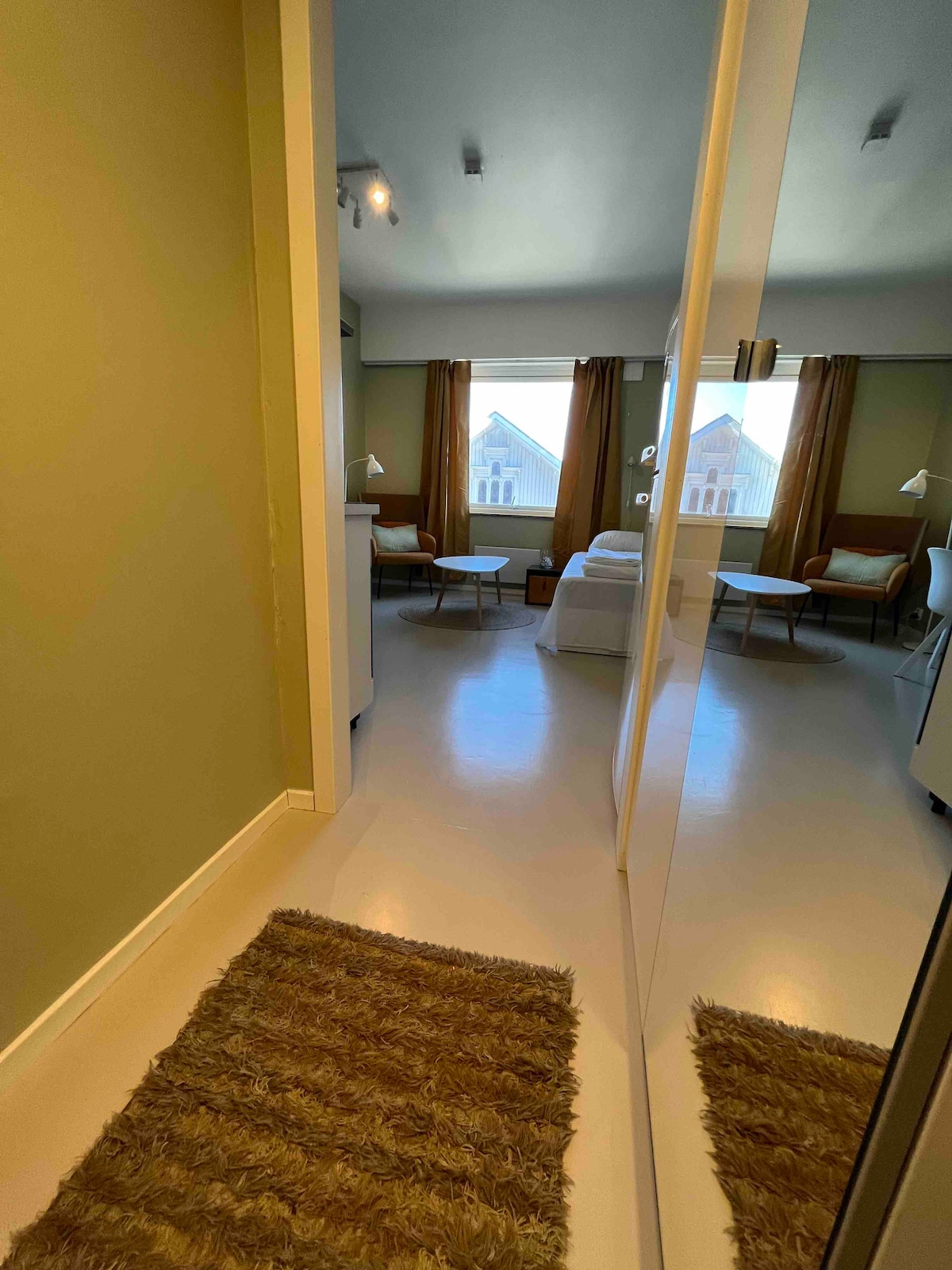 A hallway leads into a bright studio space featuring large windows with sheer curtains. Two small armchairs are positioned near a side table, and a cozy seating arrangement is visible in the background. Light-colored walls enhance the welcoming atmosphere.