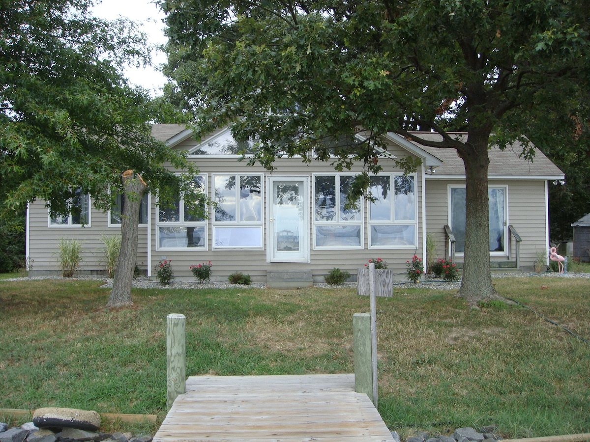 A cozy waterfront cottage is framed by mature trees, with a spacious front porch featuring large windows. A gentle slope leads to a pier, offering access to the water. The yard is landscaped with simple flower beds and a clear path from the pier to the home.
