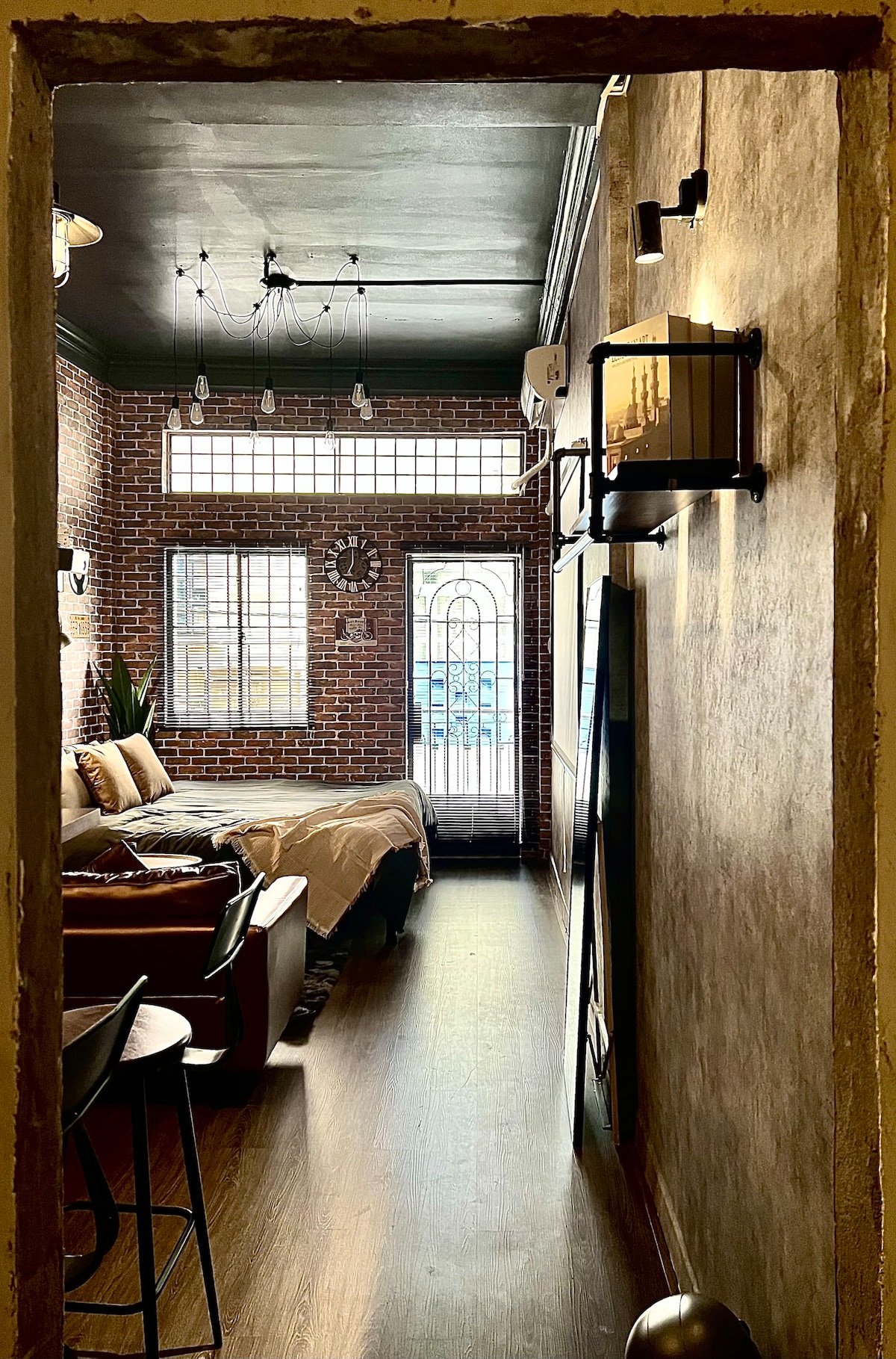 The entrance view of a studio apartment is captured, featuring a cozy sleeping area with a large bed and a sofa. Natural light filters through the large windows, illuminating the rustic brick walls. A small dining area with chairs is placed near the entrance, enhancing the inviting space.