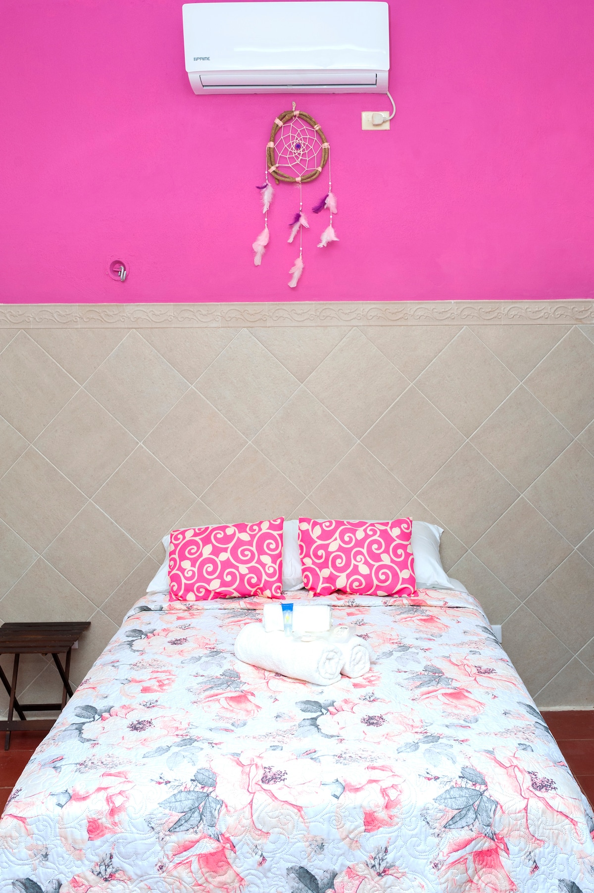 A private room is presented, featuring a spacious bed with a floral patterned quilt. Bright pink walls add color, while a dreamcatcher decorates the area above the bed. Fresh towels are neatly arranged on the bed, and a small wooden table sits beside it.