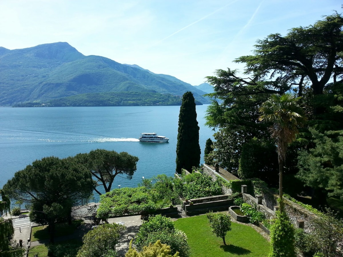 The image captures a serene view of the lake surrounded by green hills and mountains. A boat is seen gliding across the water, while lush trees and gardens are framed in the foreground, suggesting a peaceful outdoor environment.