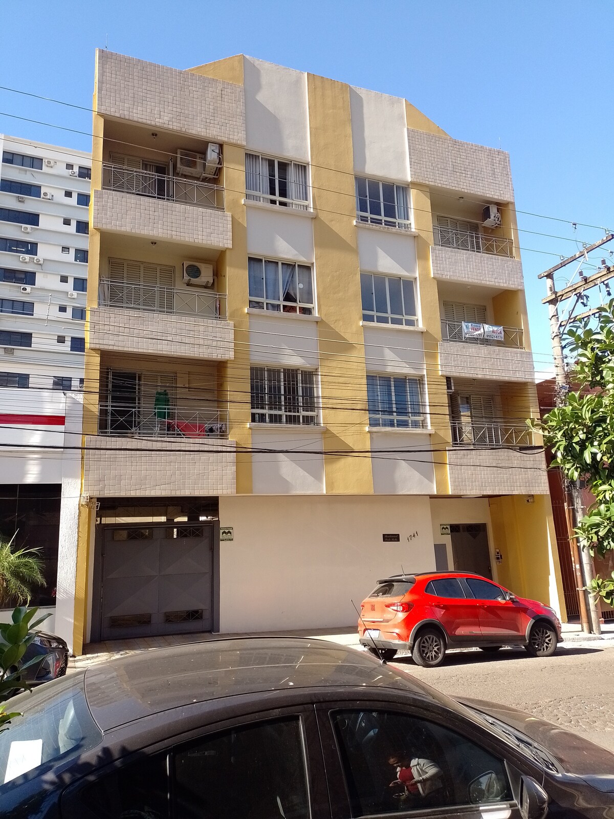 A modern, three-story building is featured with a yellow and white facade. Balconies are visible on each floor, complemented by metal railings. An orange vehicle is parked in front, with another black car partially in view. Clear blue skies provide a bright backdrop for the structure.
