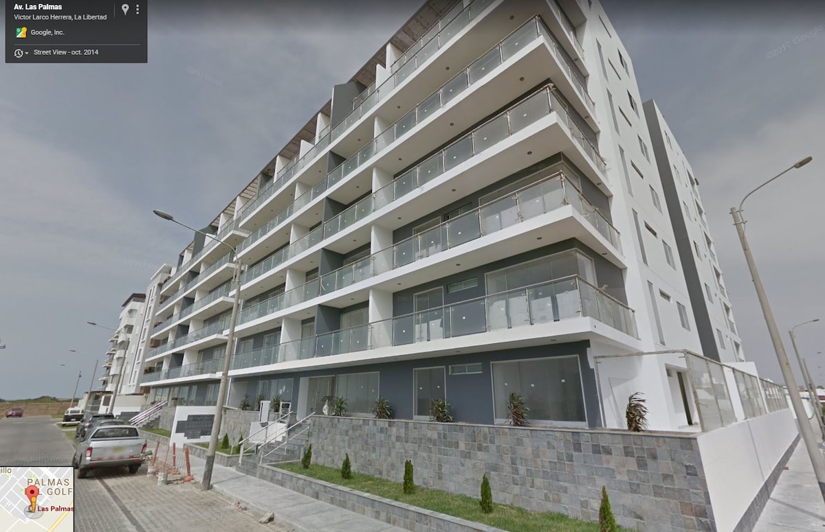 A modern building with multiple levels is showcased from the exterior, featuring large glass balconies and a sleek façade. The ground level reveals a landscaped area with neatly arranged plants, and wide pathways are visible, contributing to an open and contemporary environment.